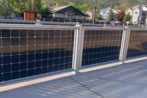 a modern solar panel fence with galvanized steel framing along a sidewalk in a suburban neighborhood with hills and houses in the background