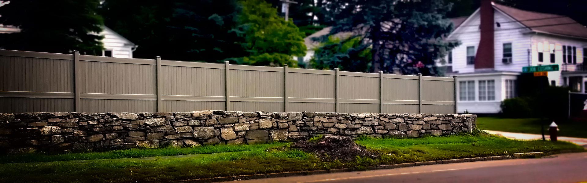 New vinyl fence installation by Miller Fence A light brown vinyl fence above stone wall along a residential street with a white house in the background